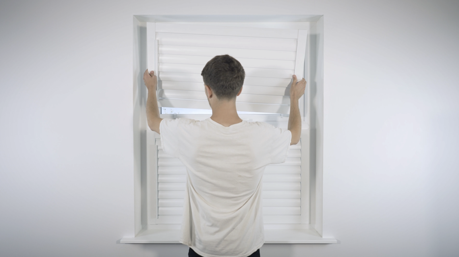 Man fitting white interior window shutters, standing at a brick-less window opening indoors.