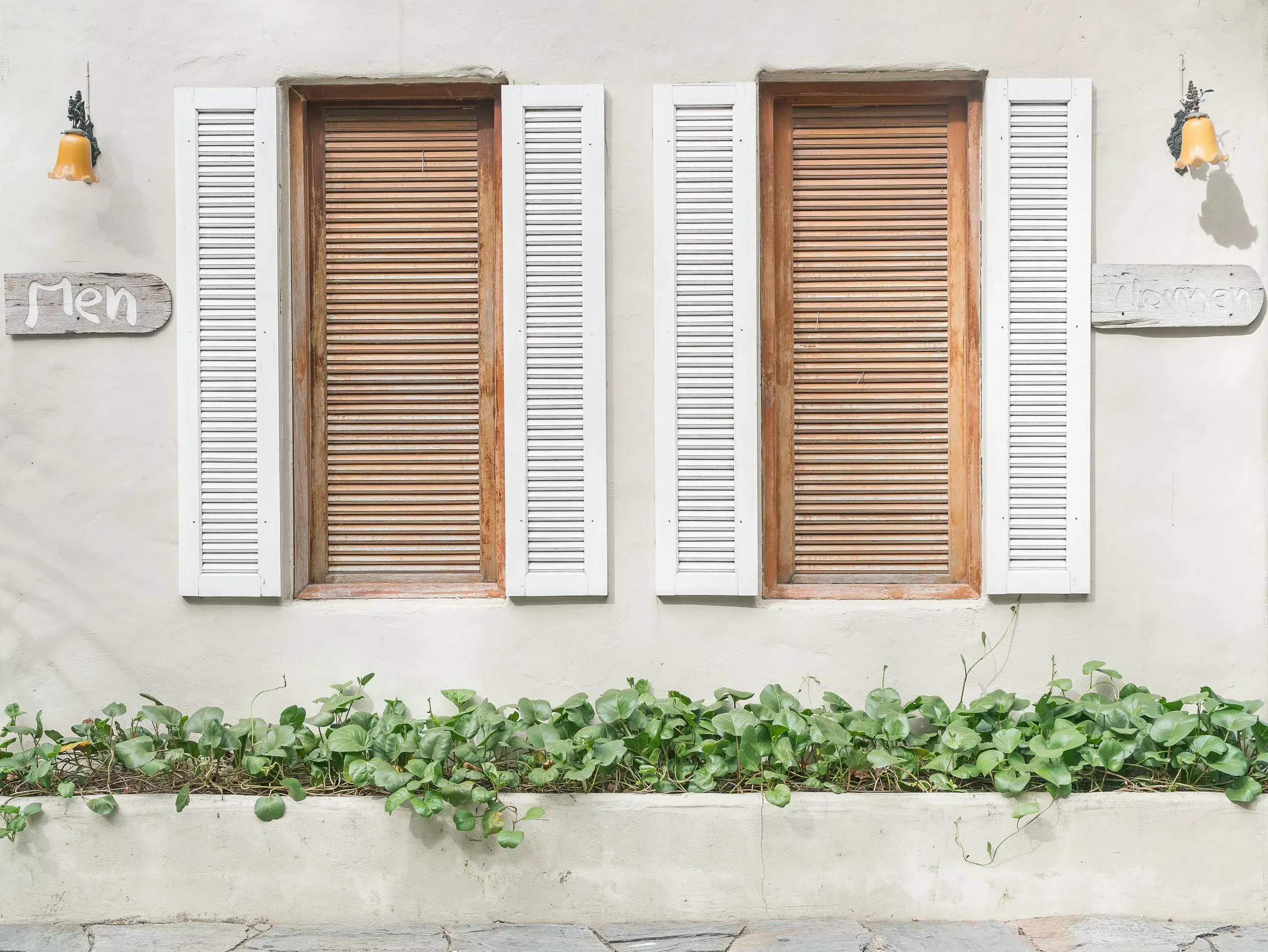 Two shuttered windows with contrasting louvred slats sit in a light wall, framed by open and closed shutters. A low planter bed with green trailing plants runs along the base. How to fit window shutters uk integrated naturally.