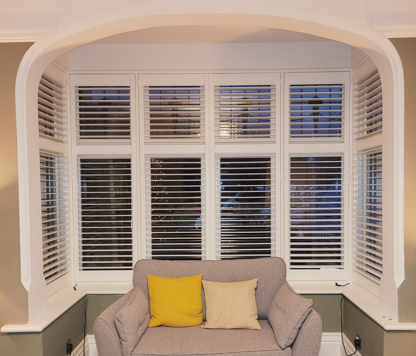 Wide bay window with white plantation shutters and arched top, built-in window seat with cushions in grey and yellow accents. How to fit window shutters UK integrated.